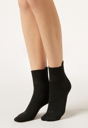 Par de calcetines negros texturizados hasta el tobillo, usados en los pies de una persona, mostrados contra un fondo claro y liso.