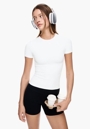 Weißes, kurzärmliges, figurbetontes Oberteil, kombiniert mit schwarzen Shorts. Das Model hält einen Kaffeebecherhalter mit zwei Tassen. Sanfte Texturen und einfache Designelemente.