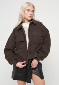 Mlada ženska z dolgimi blond lasmi, oblečena v temno rjavo jakno na gumbe, bež pulover in črno usnjeno krilo s sprednjim razporkom.