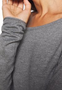 Maglia grigia a maniche lunghe con scollatura tonda, realizzata in un tessuto morbido e texturizzato e con maniche aderenti, che mostra la spalla e la mano di chi la indossa.