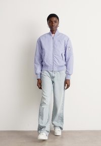 Svijetlo ljubičasta bomber jakna s prednim zatvaračem, rebrastim manžetama i bočnim džepovima. Nosena s svijetloplavim trapericama i bijelim tenisicama.