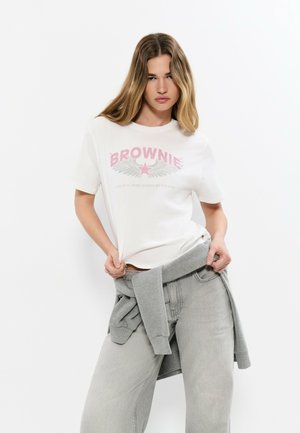 Mujer joven con cabello largo, vistiendo una camiseta blanca con el gráfico "BROWNIE", jeans gris claro y una sudadera gris atada a la cintura.