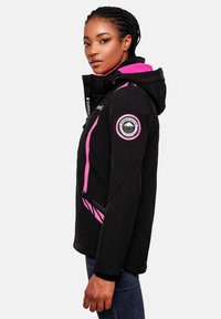 Woman in profile wearing a black and pink hooded jacket with a mountain logo patch on the sleeve, paired with blue jeans.