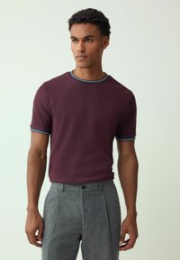 Tamsiai raudonas trumpomis rankovėmis megztinis su tekstūruotu raštu ir apvalia apykakle, su mėlynais ir baltais dryžuotais akcentais ant apykaklės ir rankovių.