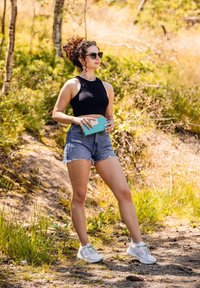 Woman wearing a black sleeveless top, denim shorts, and white sneakers, holding a turquoise clutch with a polka dot design in an outdoor setting.