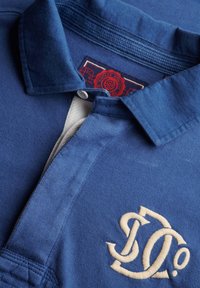 Blauw poloshirt met button-down kraag, wit geborduurd "SD"-logo op de borst en een rood vintage atletieklabel aan de binnenkant van de kraag.