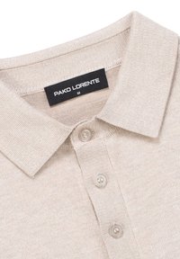 Beżowa dzianinowa koszulka polo z trzema guzikami i czarną metką z napisem "PAKO LORENTE M" przy kołnierzyku.