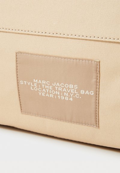 Beige kangast reisikott, millel on ristkülikukujuline silt, millel on valge tekst, mis ütleb: "MARC JACOBS, STiIL: REISIKOTT, ASUKOHT: N.Y.C., AASTA: 1984."
