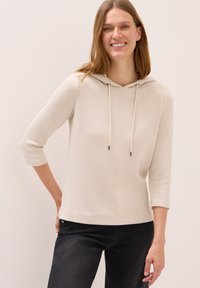 Kvinna som ler i en ljusbeige ribbad hoodie med trekvartslånga ärmar och svarta jeans, stående mot en enkel ljus bakgrund.