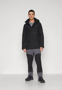 Musta vedenpitävä takki, jossa on vetoketjullinen etuosa, kaksi rintataskua ja huppu; yhdistetty harmaisiin ja mustiin urheiluhousuihin sekä mustiin lenkkareihin.