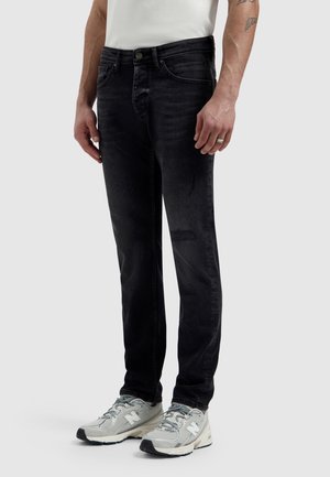 Man draagt vervaagde zwarte spijkerbroek, witte sneakers en een wit shirt, staand tegen een effen lichtgrijze achtergrond.