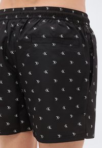 Μαύρα μαγιό σορτς με ελαστική μέση, διακοσμημένα με επαναλαμβανόμενο λευκό λογότυπο και πίσω τσέπη. Λείο, ελαφρύ υλικό.