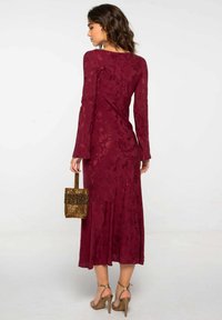 Vestido de encaje burdeos con mangas largas y un dobladillo acampanado, acompañado de un bolso dorado con flecos, sobre un fondo liso.