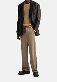 Blazer en cuir noir, pull en tricot beige et pantalon marron à carreaux avec cordon de serrage. Accents marron sur le côté du pantalon. Chaussures noires épaisses.