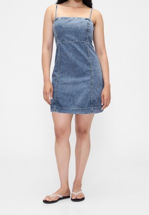 Kvinde iført en blå denim mini kjole med tynde stropper og sorte klipklappere, stående mod en ensfarvet hvid baggrund.