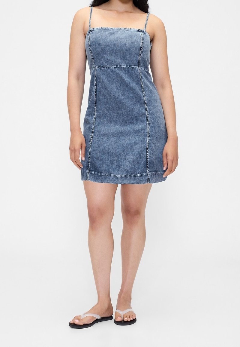 Femme portant une mini-robe en jean bleu à fines bretelles et des tongs noires, debout sur un fond blanc uni.