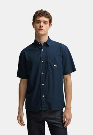 Camicia a maniche corte di colore blu navy con bottone, realizzata in tessuto liscio, dotata di taschino sul petto con un logo a contrasto in rosso, bianco e blu.
