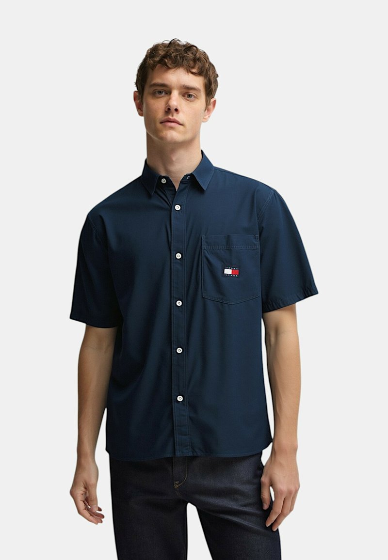 Camicia a maniche corte di colore blu navy con bottone, realizzata in tessuto liscio, dotata di taschino sul petto con un logo a contrasto in rosso, bianco e blu.