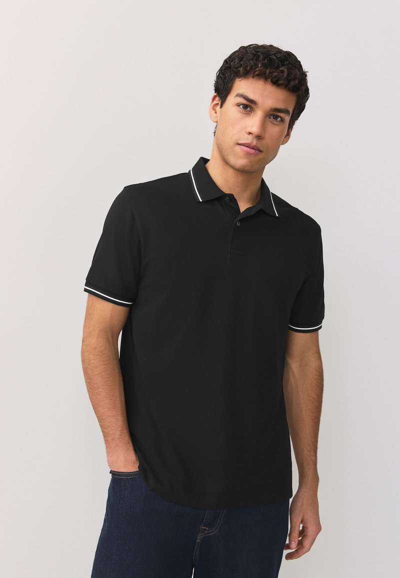 Jeune homme debout, une main dans la poche, portant un polo noir avec des bordures blanches au col et aux manches, et un jean bleu foncé.