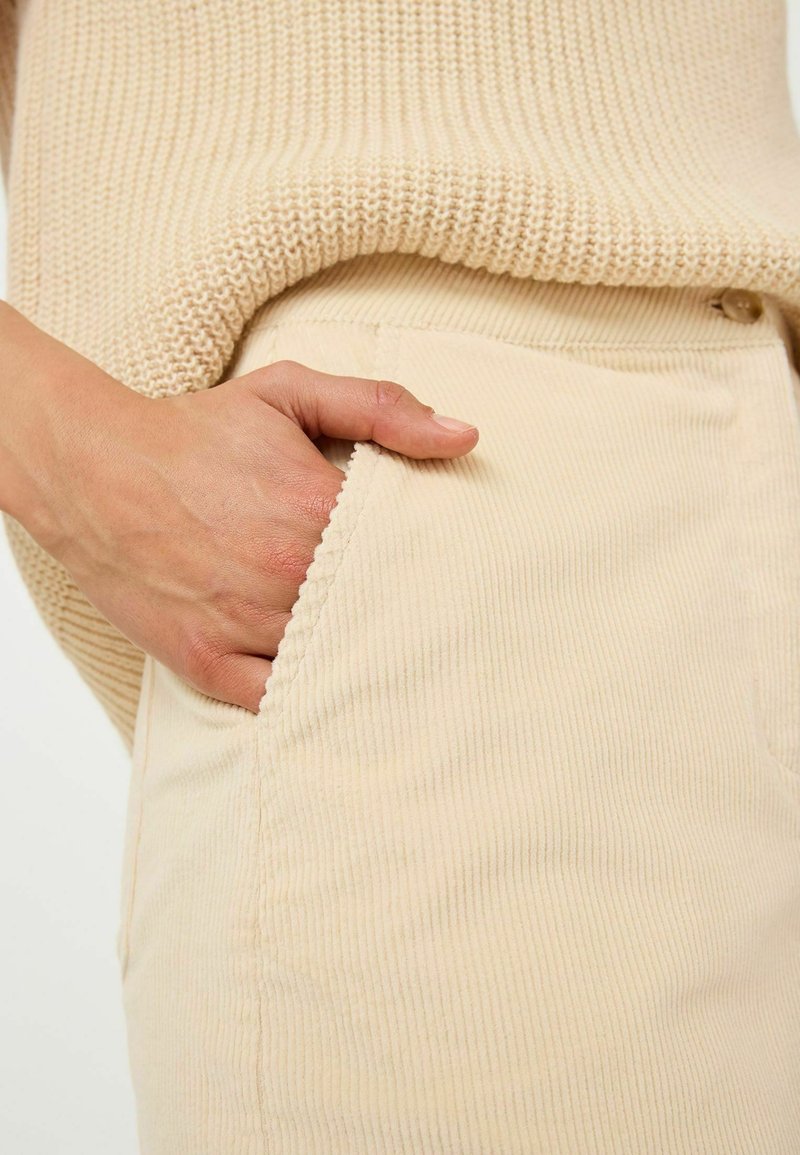 Pantalon en velours côtelé crème avec une poche latérale, associé à un pull beige doux et texturé. Main reposant calmement dans la poche.