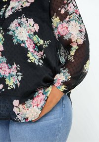 Blouse noire transparente à motif floral rose, blanc et jaune, portée avec un jean denim bleu clair, main dans la poche.