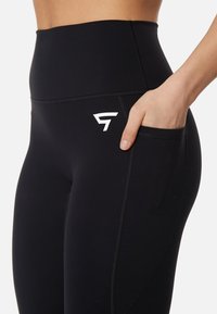 Leggings nere a vita alta realizzati in tessuto elasticizzato, dotati di una tasca laterale e di un logo bianco sulla coscia superiore. Texture liscia, design elegante.