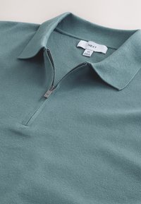 Grünes, gestricktes Poloshirt mit einem Reißverschluss am Hals. Verfügt über eine glatte Textur und einen klassischen Kragen. Etikett innen am Kragen sichtbar. Größe M.