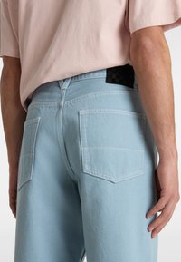 Light blue denim jeans with a relaxed fit, featuring two back pockets and a black checkerboard patch on the waistband.