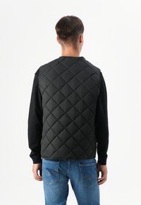 Mladog muškarca vidimo s leđa, nosi crnu jaku quilted prsluk preko dugih rukava košulje i plavih traperica, na jednostavnoj bijeloj pozadini.
