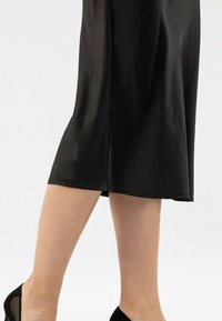 Abito nero di lunghezza al ginocchio con una texture satinata liscia, caratterizzato da un orlo dritto e un drappeggio sottile, abbinato a scarpe nere con tacco alto.