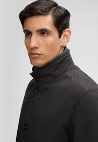 Cappotto nero con colletto alto, caratterizzato da due grandi bottoni, texture liscia e design su misura, che presenta una silhouette moderna e strutturata.