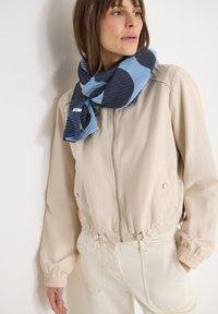 Femme portant une veste beige zippée, un pantalon crème et une écharpe à motif bleu texturée, debout contre un mur blanc, regardant vers la droite.