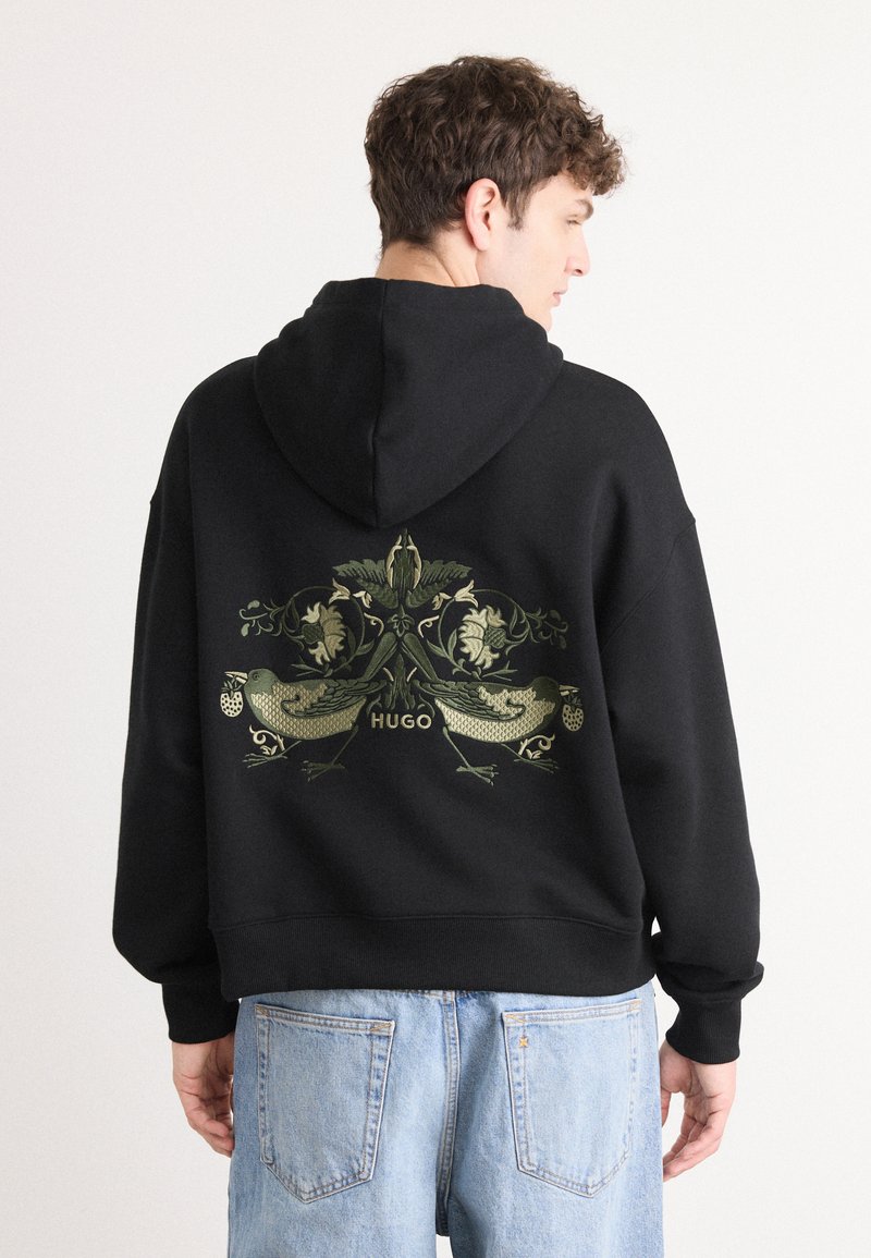 Schwarzer Hoodie mit einem großen grünen Blumen- und Vogelmotiv auf der Rückseite. Er besitzt eine Kapuze mit Kordelzug und gerippte Bündchen und wird mit blauen Jeans kombiniert.