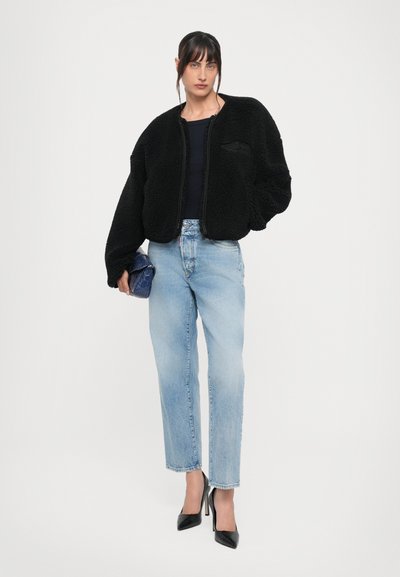 Veste en fausse fourrure noire avec fermeture éclair à l'avant, associée à un jean droit bleu clair et des escarpins noirs. Petit sac à main bleu à la main.