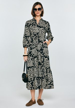 Kobieta w czarno-białej sukience midi w wzór paisley z rękawami 3/4, brązowymi butami, czarną torebką, masywnymi bransoletkami i dużymi okularami przeciwsłonecznymi.