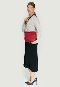 Bolso bandolera de cuero rojo con acabado texturizado, cierre de cremallera y correa ajustable, combinado con un suéter ligero y una falda negra.