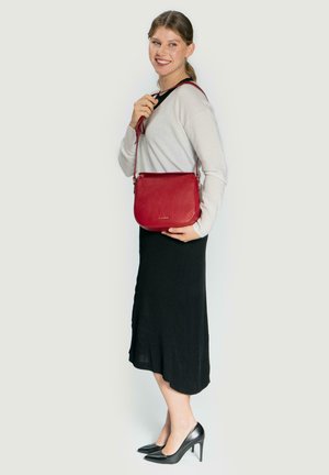 Rote Leder-Umhängetasche mit strukturiertem Finish, Reißverschluss und verstellbarem Trageriemen, kombiniert mit einem leichten Pullover und einem schwarzen Rock.