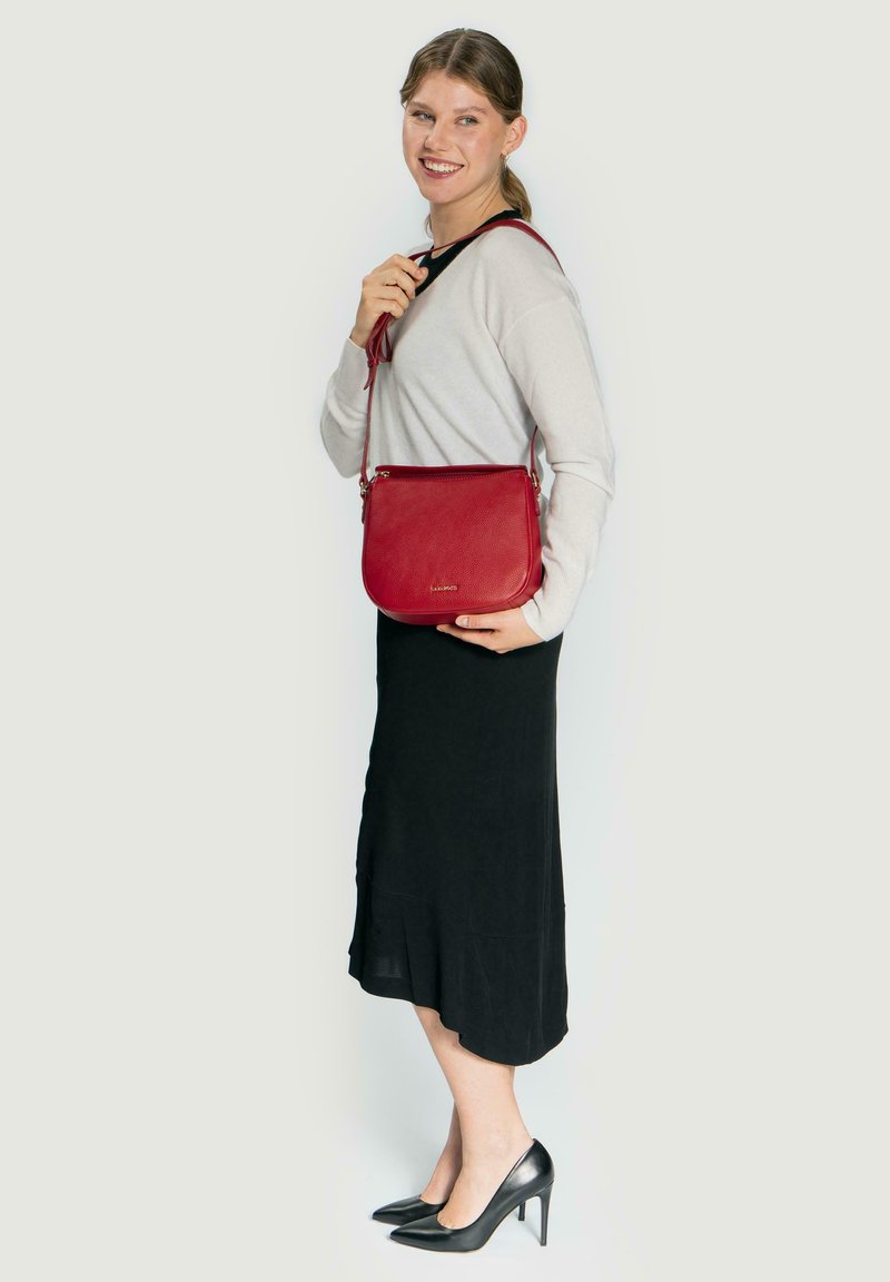 Bolso bandolera de cuero rojo con acabado texturizado, cierre de cremallera y correa ajustable, combinado con un suéter ligero y una falda negra.