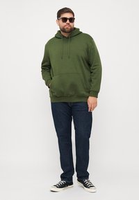 Olive green hoodie with front pocket, adjustable drawstring, and ribbed cuffs; paired with dark blue jeans and black canvas sneakers.