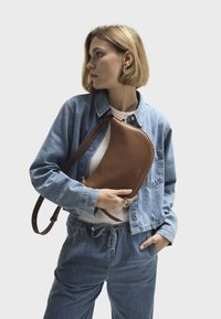 Brun læder crossover-taske med en buet form og skulderrem, holdt mod en denimjakke og en lys farvet top, der fremhæver det enkle design.