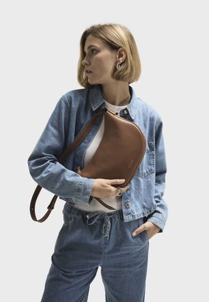 Brown leather crossbody bag with a curved shape and shoulder strap, held against a denim jacket and light-colored top, showcasing simple design.