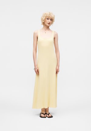 Γυναίκα με κοντά σγουρά ξανθά μαλλιά που φοράει αμάνικο ανοιχτό κίτρινο μαξι φόρεμα και μαύρα σανδάλια, όρθια μπροστά σε απλό φόντο.