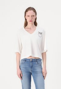 T-shirt bianco cropped a maniche corte con scollatura a V, decorato con una grafica di due uccelli all'interno di un cerchio nella zona del petto sinistro.