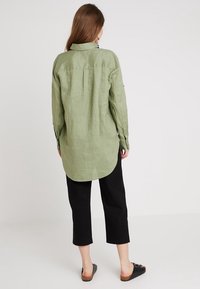 Koszula z długim rękawem w jasnym oliwkowym kolorze, o prostym kroju, z kołnierzykiem i zakładką z tyłu, zestawiona z czarnymi, krótkimi spodniami i czarnymi sandałami na płaskiej podeszwie.