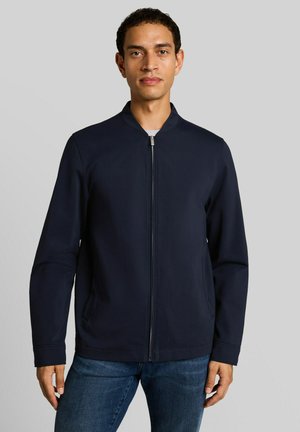 Uomo che indossa una giacca blu navy con zip e collo alto e jeans blu scuro, in piedi davanti a uno sfondo chiaro e uniforme.
