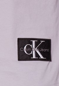 Světle fialová bavlněná látka s černě vyšívanou nášivkou s textem "Calvin Klein Jeans" a bílým logem "CK". Hladká textura, obdélníková nášivka.