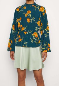 Blommig långärmad topp i teal med gula och orange blommor, rynkad halsringning och peplummidja, kombinerad med en mintgrön satinkjol.