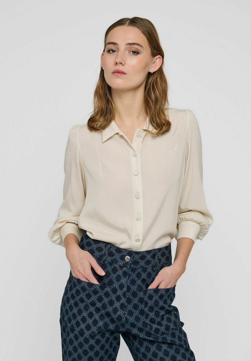 Femme aux cheveux mi-longs portant une chemise crème boutonnée et un pantalon sombre à motifs, debout les mains dans les poches devant un fond uni.