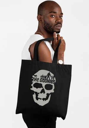 Mężczyzna z brodą i białą koszulką bez rękawów trzyma czarną torbę na ramię z dużą grafiką czaszki i napisem „The Goonies”.