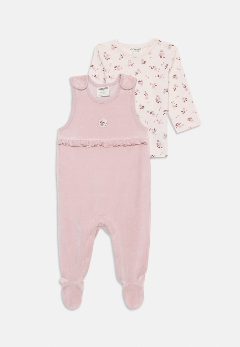 Rosa velour overall med volangkant og knappedetaljer, kombinert med en langermet topp med blomsterdesign på lys bakgrunn.